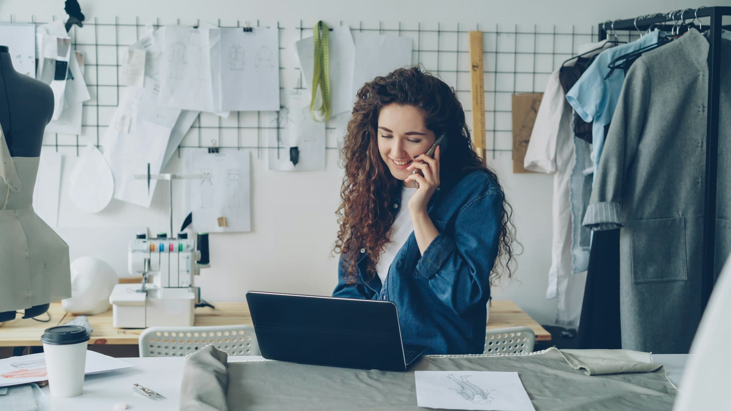A designer on the phone at her workbench, laptop open